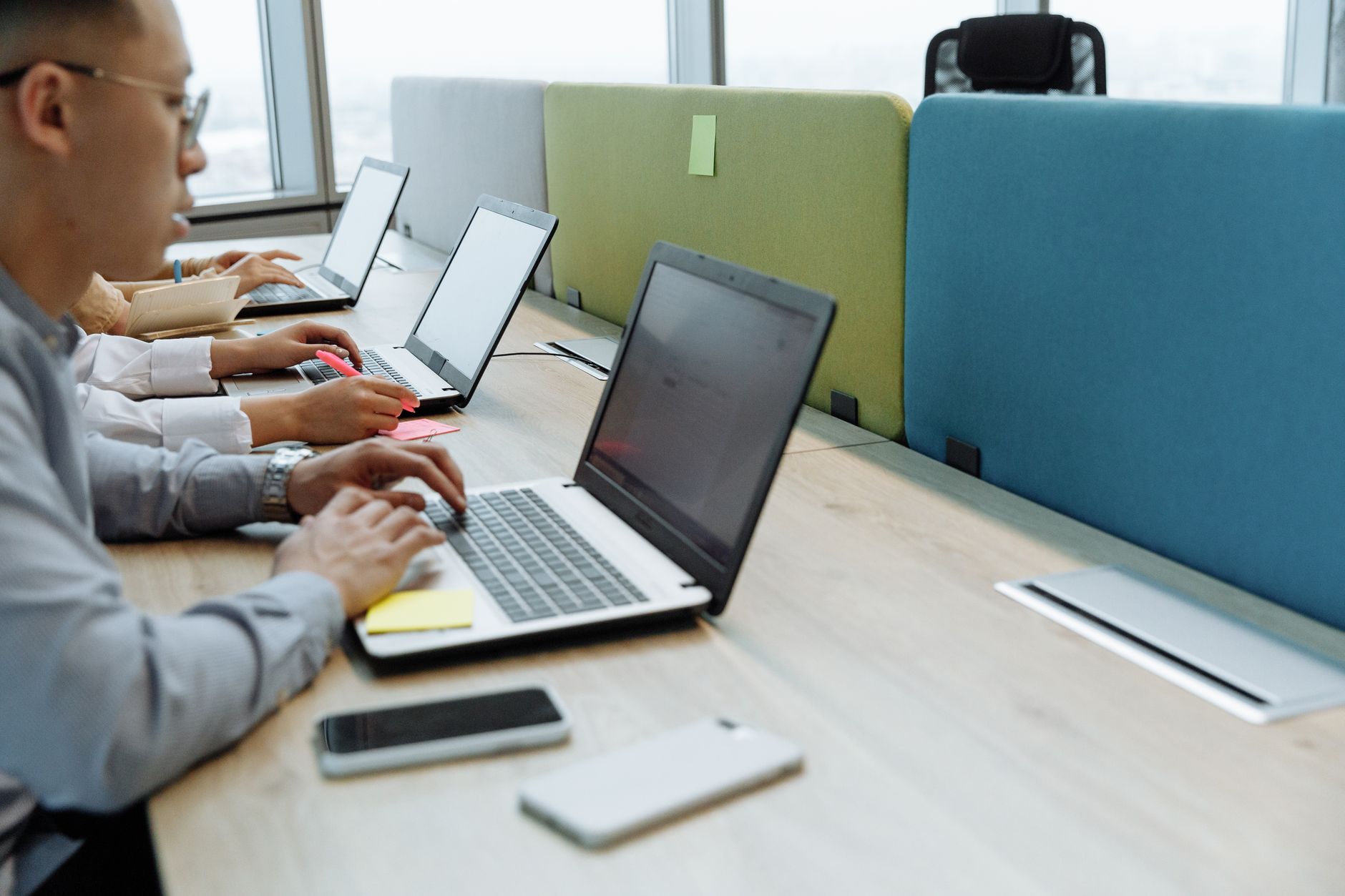 Des personnes travaillent sur des ordinateurs portables le long d’une longue table de bureau partagé, avec des cloisons vertes et bleues entre les postes.
