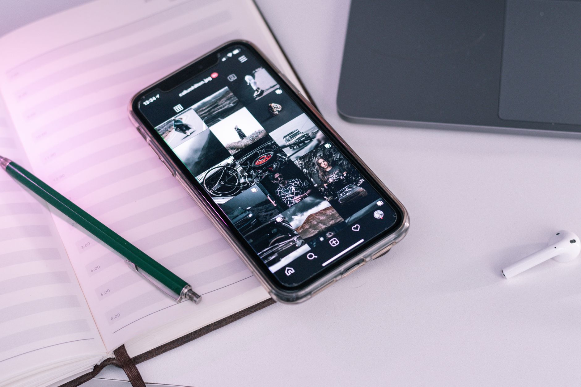 Smartphone affichant une grille de photos sur un carnet ouvert, avec un stylo vert, un ordinateur portable et un écouteur sans fil sur un bureau clair.